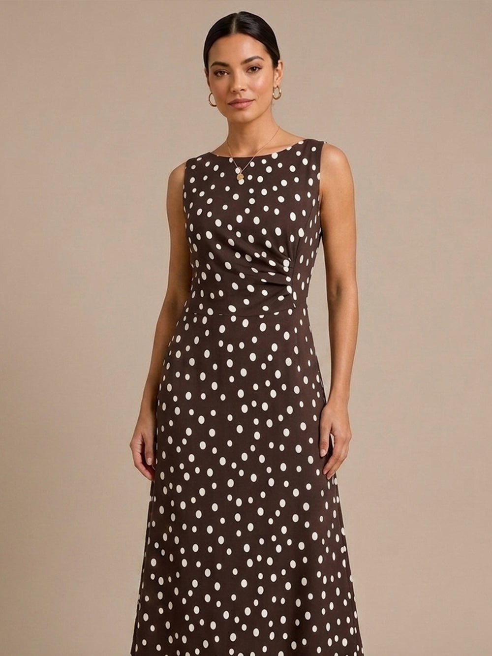 A woman modeling a sleeveless brown maxi dress with a white polka dot pattern against a solid beige studio background.